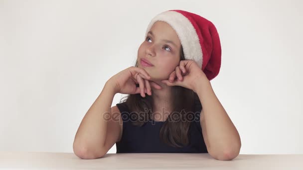 Menina adolescente bonita em Santa Claus chapéu sentado e sonhando com um presente, expressa felicidade e antecipação no fundo branco imagens de vídeo — Vídeo de Stock