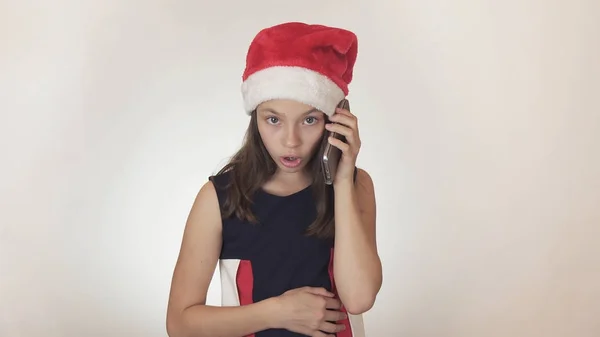Hermosa chica traviesa adolescente en un sombrero de Santa Claus se comunica emocionalmente en el teléfono inteligente sobre fondo blanco —  Fotos de Stock
