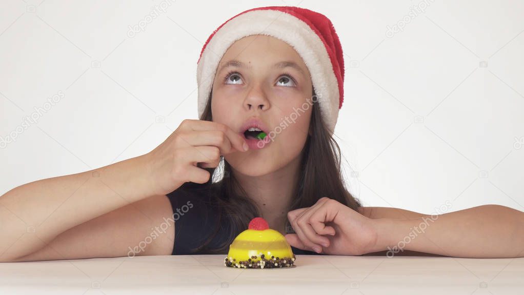 Beautiful teen girl in Santa Claus hat with an appetite and pleasure eating a birthday cake on white background