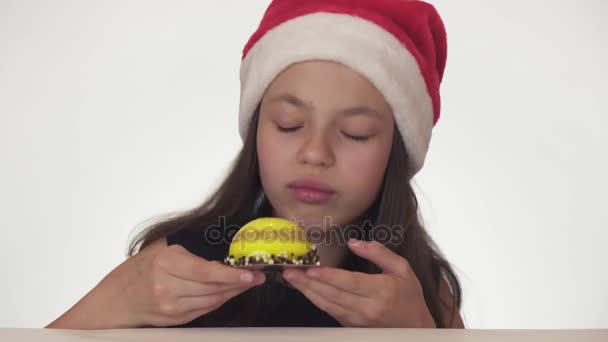 Όμορφη έφηβος κορίτσι στον Αϊ Βασίλη καπέλο με όρεξη και χαρά που τρώει μια τούρτα γενεθλίων σε λευκό φόντο πλάνα βίντεο — Αρχείο Βίντεο