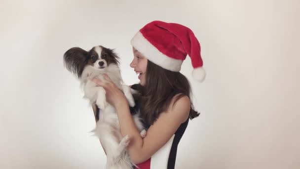 Bir Noel Baba şapkası ve sevinçle öpüşme ve beyaz arka plan stok görüntüleri video dalga geçmeyi köpek kıta oyuncak İspanyol Papillon güzel genç kız. — Stok video