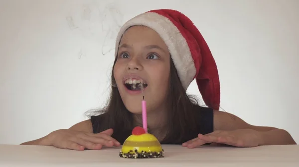 Krásná zlobivá holka teenager v Santa Claus klobouk sfoukne svíčku na sváteční dort na bílém pozadí — Stock fotografie