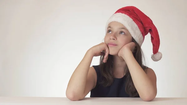 Hermosa chica adolescente en Santa Claus sombrero sentado y soñando con un regalo, expresa la felicidad y la anticipación en el fondo blanco —  Fotos de Stock