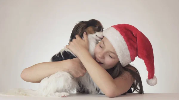 Hermosa adolescente en Santa Claus sombrero felizmente abraza a su perro sobre fondo blanco —  Fotos de Stock