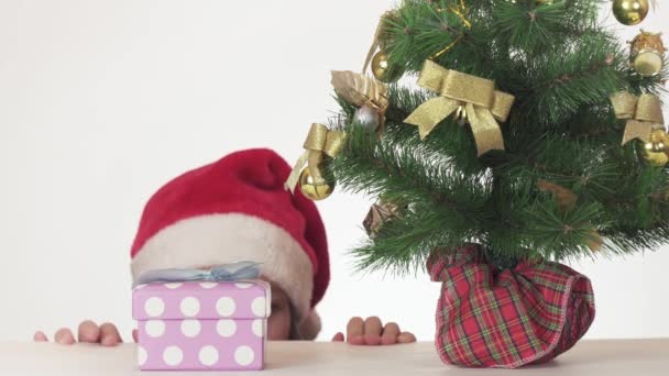 Hermosa chica traviesa adolescente en un sombrero de Santa Claus abre secretamente un regalo bajo el árbol de Año Nuevo en el video de imágenes de fondo blanco . — Vídeos de Stock