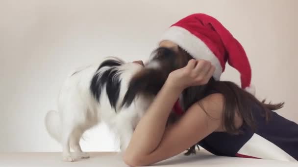 Mooie tiener meisje en hond continentaal Toy Spaniel Papillon in Santa Claus kappen vreugdevol zoenen en gek rond op witte achtergrond stock footage video — Stockvideo