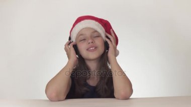 Güzel yaramaz kız genç bir Noel Baba şapkalı kulaklıklar müzik dinler ve boyunca üzerinde beyaz arka plan stok görüntüleri video şarkı söylüyor.