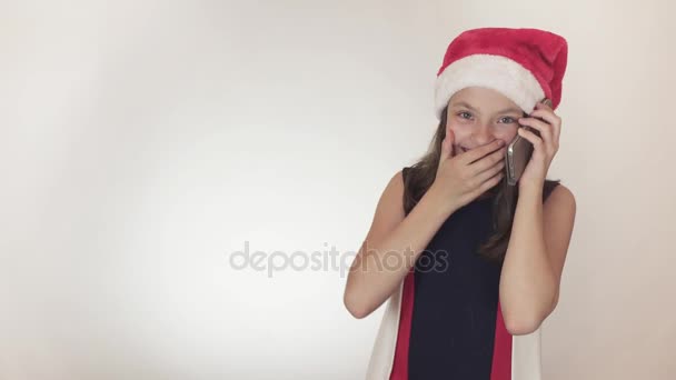 Hermosa chica traviesa adolescente en un sombrero de Santa Claus se comunica emocionalmente en el teléfono inteligente en el video de imágenes de fondo blanco . — Vídeos de Stock