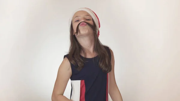 Beautiful happy girl teenager in a Santa Claus hat emotionally fools around and makes a mustache out of her hair on white background — Stock Photo, Image