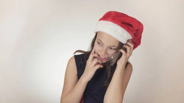 Linda menina safada adolescente em um chapéu de Papai Noel se comunica emocionalmente no smartphone no fundo branco — Fotografia de Stock