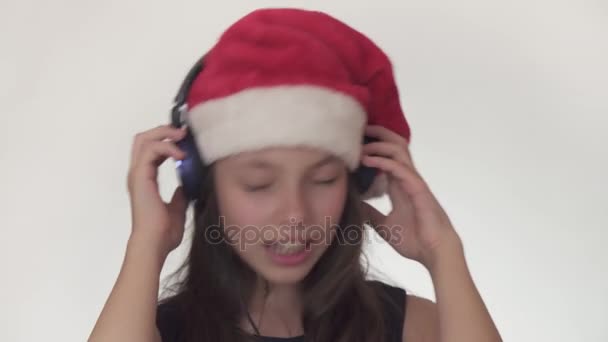Güzel yaramaz kız genç bir Noel Baba şapkalı kulaklıklar müzik dinler ve boyunca üzerinde beyaz arka plan stok görüntüleri video şarkı söylüyor. — Stok video