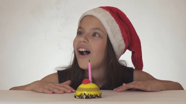 Beautiful naughty girl teenager in a Santa Claus hat blows out a candle on a festive cake on white background — Stock Photo, Image