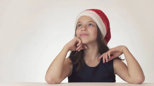 Bella ragazza adolescente in cappello di Babbo Natale seduto e sognando un regalo, esprime felicità e anticipazione su sfondo bianco — Foto Stock