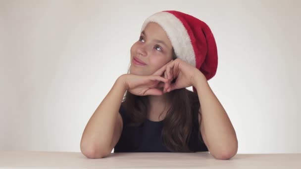 Vackra glada teen flicka i en jultomten hatt drömmer om en gåva, får en festlig tårta och uttrycker missnöje på vit bakgrund arkivfilmer video — Stockvideo