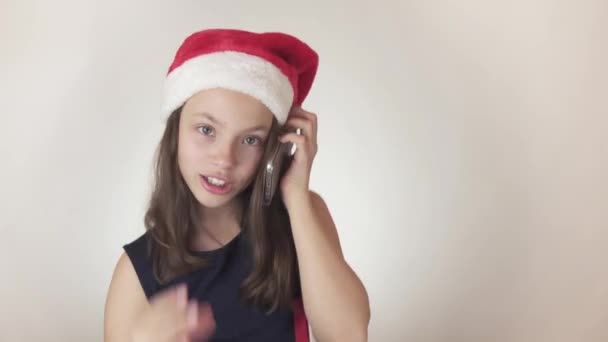 Krásná zlobivá holka teenager v Santa Claus klobouk citově komunikuje na smartphone na bílém pozadí stopáže videa. — Stock video