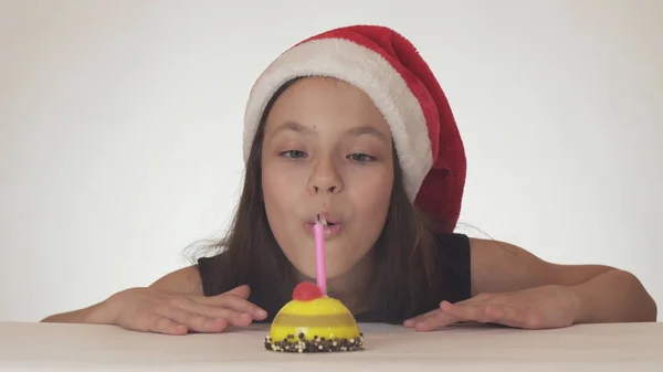 Krásná zlobivá holka teenager v Santa Claus klobouk sfoukne svíčku na sváteční dort na bílém pozadí — Stock fotografie