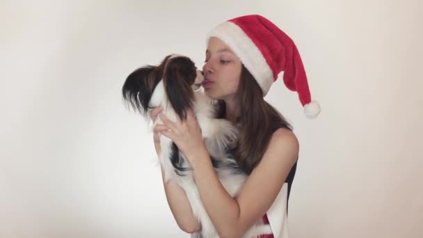 Hermosa chica adolescente en una gorra de Santa Claus y perro Continental Juguete Spaniel Papillon alegremente besando y engañando sobre fondo blanco material de archivo de vídeo . — Vídeos de Stock