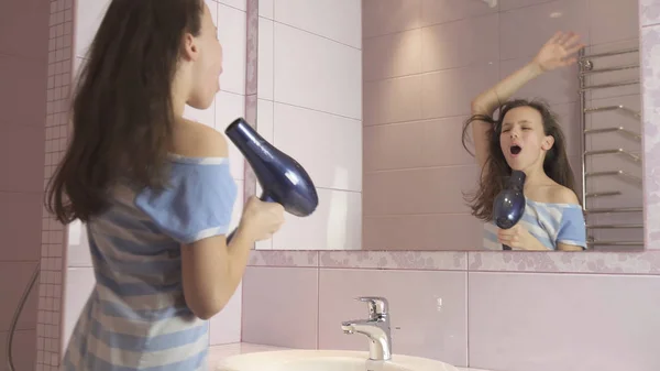 Szép boldog lány tinédzser szárítja a haját hajszárítóval és énekel és táncol a fürdőszobában tükör előtt — Stock Fotó