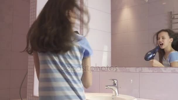 Krásná šťastná dívka teenager vysouší vlasy s vysoušečem vlasů a zpívá a tancuje před zrcadlem v koupelně stopáže videa — Stock video