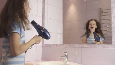 Güzel mutlu kız genç saç kurutma makinesi ile saç kurur ve şarkı söyler ve banyoda bir ayna önünde dans ediyor