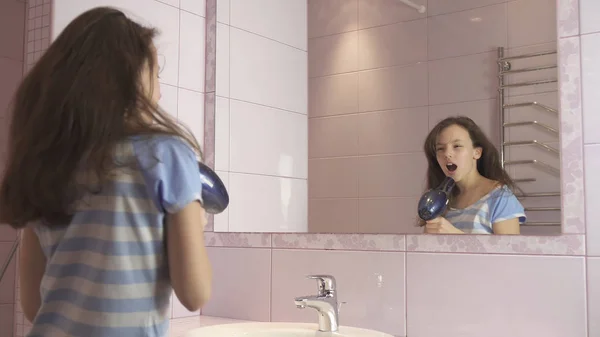 Szép boldog lány tinédzser szárítja a haját hajszárítóval és énekel és táncol a fürdőszobában tükör előtt — Stock Fotó