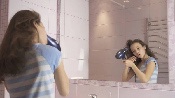 Beautiful happy girl teenager dries hair with hair dryer and sings and dances in front of a mirror in the bathroom — Stock Photo, Image