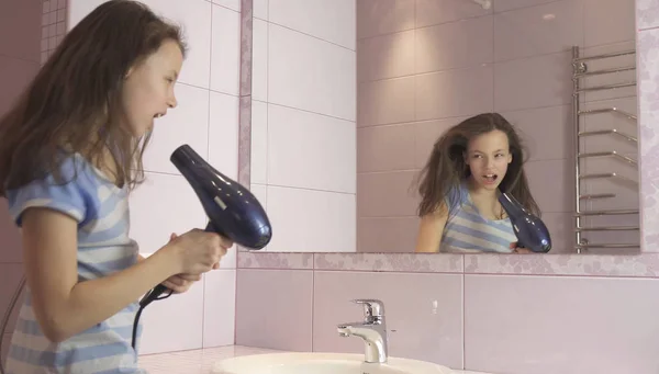 Menina feliz bonita adolescente seca o cabelo com secador de cabelo e canta e dança na frente de um espelho no banheiro — Fotografia de Stock