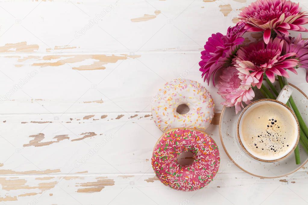 Coffee cup, donuts and gerbera flowers on white wooden table. Top view with space for your text