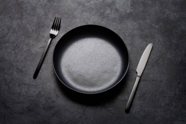 Boş tabak, çatal ve bıçak. Siyah mutfak gereçleri taş masaya dizilmiş.