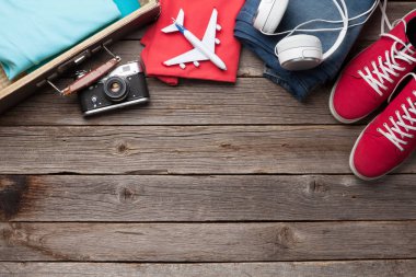 Giysiler ve seyahat aksesuarlarıyla dolu bavul. Spor ayakkabı, kot pantolon, kulaklık ve kamera. Turist ve tatil şeyleri ahşap arka planda, mesajın için yer var. Üst görünüm düzlüğü
