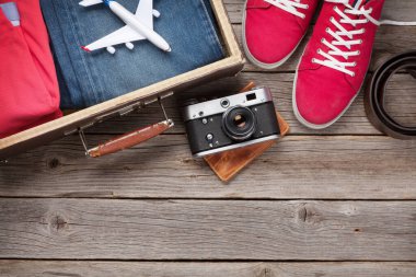 Bavul, kamera, kıyafetler ve seyahat aksesuarları. Spor ayakkabı, kot pantolon, uçak ve kamera. Turist ve tatil şeyleri ahşap arka planda, mesajın için yer var. Üst görünüm düzlüğü