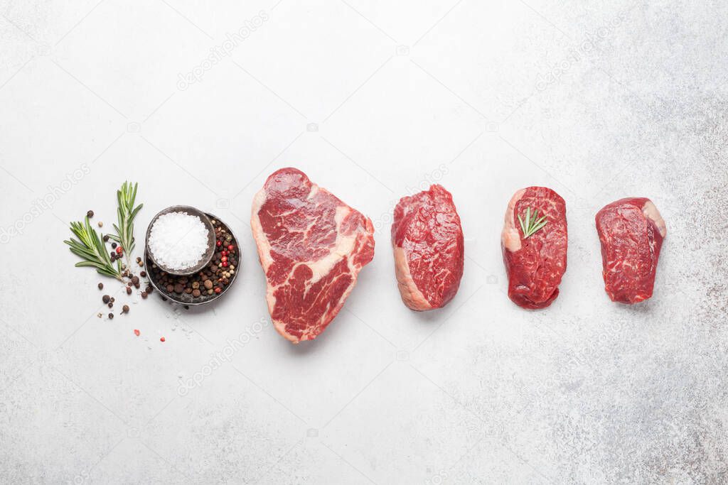 Variety of fresh raw beef steaks with spices on stone background. Top view flat lay with copy space