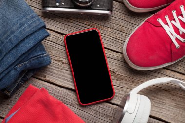 Giysiler ve aksesuarlar. Spor ayakkabı, kot pantolon, kulaklık ve akıllı telefon. Her gün için şehir kıyafeti ya da telefon ekranında fotokopi alanı olan ahşap arka planda seyahat tatili. Üst görünüm düzlüğü