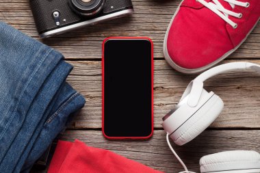 Giysiler ve aksesuarlar. Spor ayakkabı, kot pantolon, kulaklık ve akıllı telefon. Her gün için şehir kıyafeti ya da telefon ekranında fotokopi alanı olan ahşap arka planda seyahat tatili. Üst görünüm düzlüğü