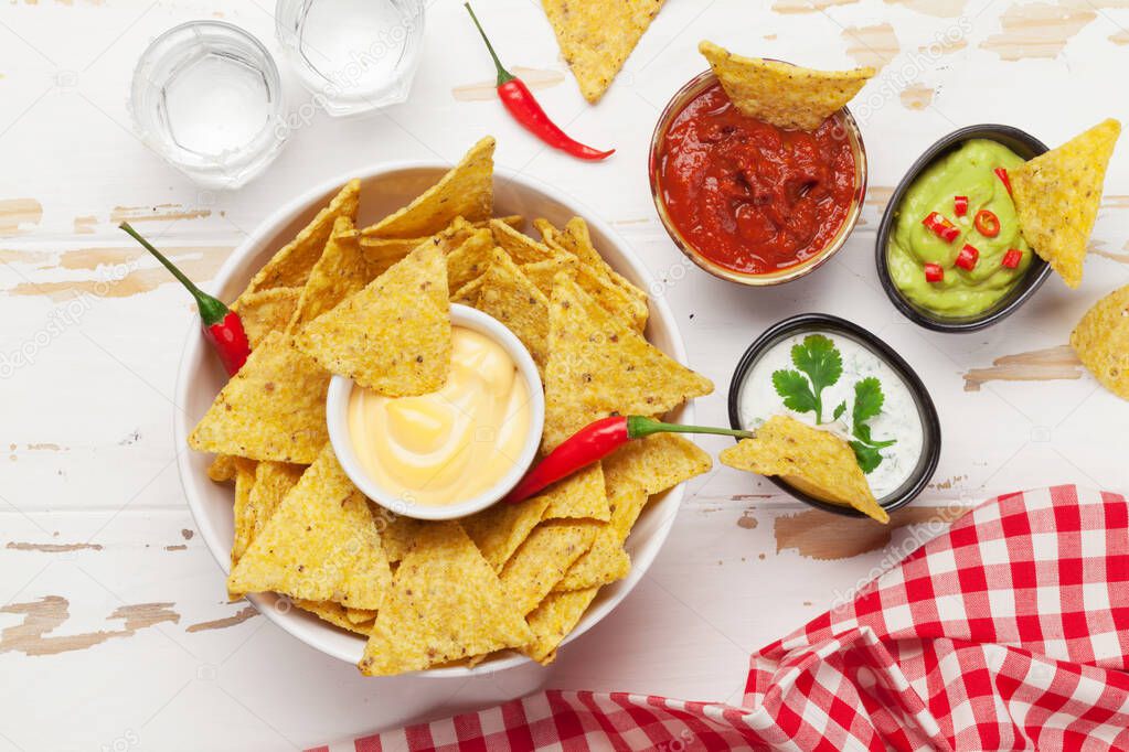 Mexican nachos chips with various sauces - guacamole, salsa, cheese and sour cream. Top view flat lay on wooden table with two tequila shots