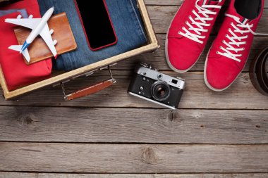Bavul, kamera, kıyafetler ve seyahat aksesuarları. Spor ayakkabı, kot pantolon, akıllı telefon ve uçak. Turist ve tatil şeyleri ahşap arka planda, mesajın için yer var. Üst görünüm düzlüğü