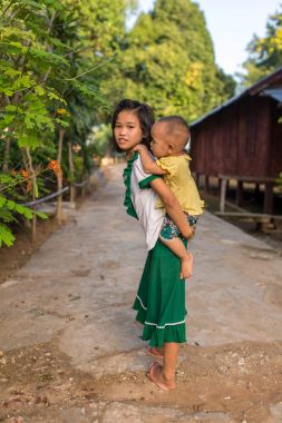 Hsipaw, Myanmar - October 7, 2016: Cute burmese girl carrying small boy in rural area in Burma. clipart