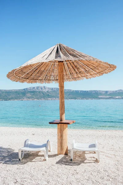 Nyári szalma esernyő és két napozóágy az üres strandon Horvátországban. — Stock Fotó