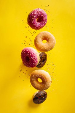Çeşitli dekore edilmiş donutlar, hareket halinde serpiştirilmiş, sarımsı arka plana dökülüyor.