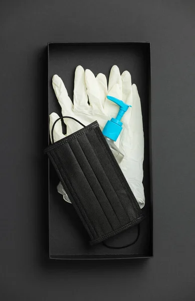 Protective face mask, sanitizer gel and lab gloves in black box on black paper background flat lay — Stock Photo, Image