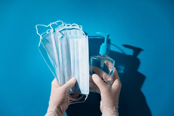 Woman hands in latex gloves holding facial medical masks and sanitizer, blue background — Stock Photo, Image