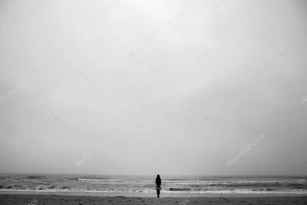 Photo of young woman with long hair standing back on beach of the North Sea in cloudy evening.