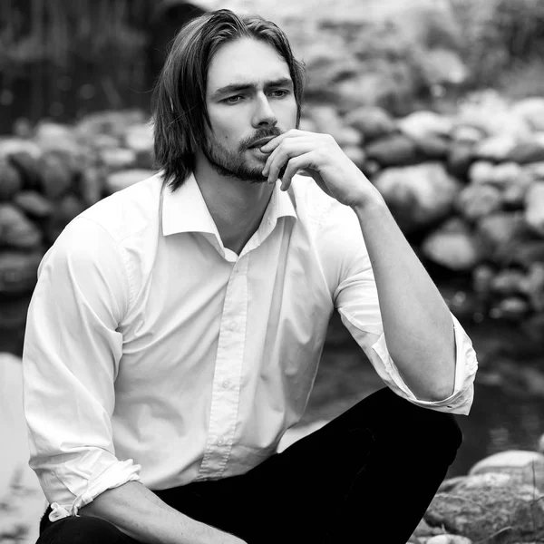 Black-white outdoor portrait of elegant long hair handsome man siting near pond. — Stock Photo, Image