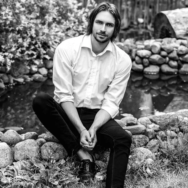 Black-white outdoor portrait of elegant long hair handsome man siting near pond. — Stock Photo, Image