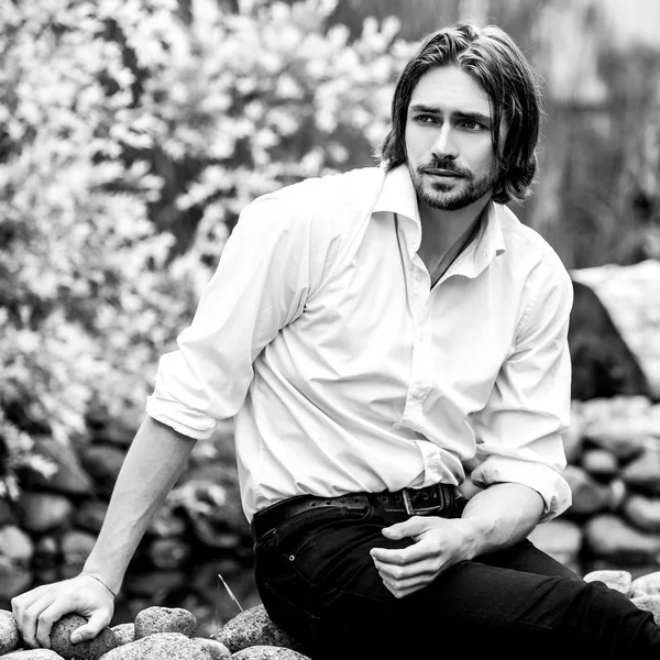 Black-white outdoor portrait of elegant long hair handsome man siting near pond. — Stock Photo, Image
