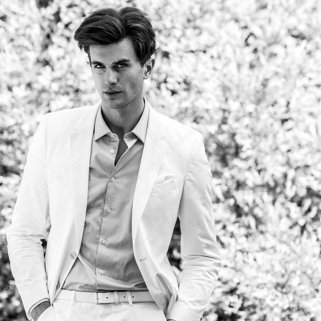 Black-white portrait of young handsome fashionable man in white suit against nature background.