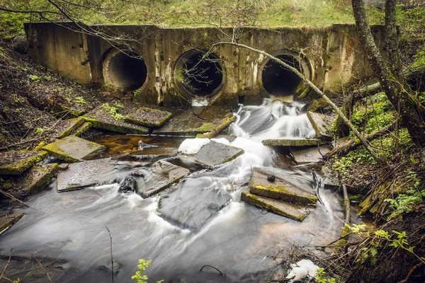 Barrage de rivière fait de tuyaux sous un pont dans une route forestière avec de l'eau courante . — Photo