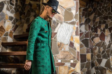 Şık yakışıklı genç uzun yeşil palto ve şapka taş arka plan.