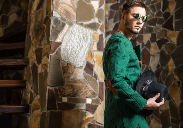 Bello giovane uomo elegante in lungo cappotto verde e cappello sullo sfondo di pietra . — Foto Stock