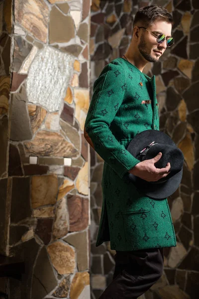 Bello giovane uomo elegante in lungo cappotto verde e cappello sullo sfondo di pietra . — Foto Stock